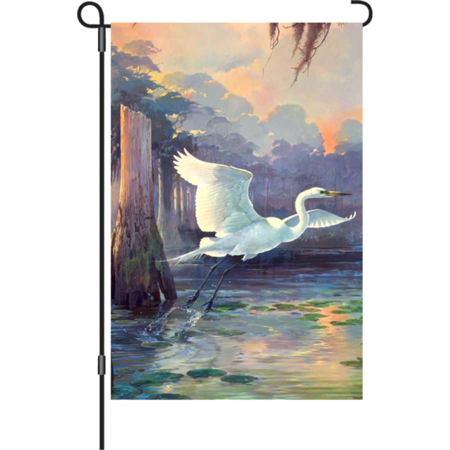 12 in. Flag - Heron Sunrise featuring a colorful heron silhouette against a sunrise, displayed in an outdoor garden setting.