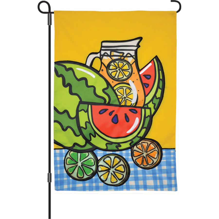 12 in. Watermelon Citrus garden flag featuring a playful design with watermelon and citrus fruits, displayed on a sturdy flag stand in a sunny outdoor setting.
