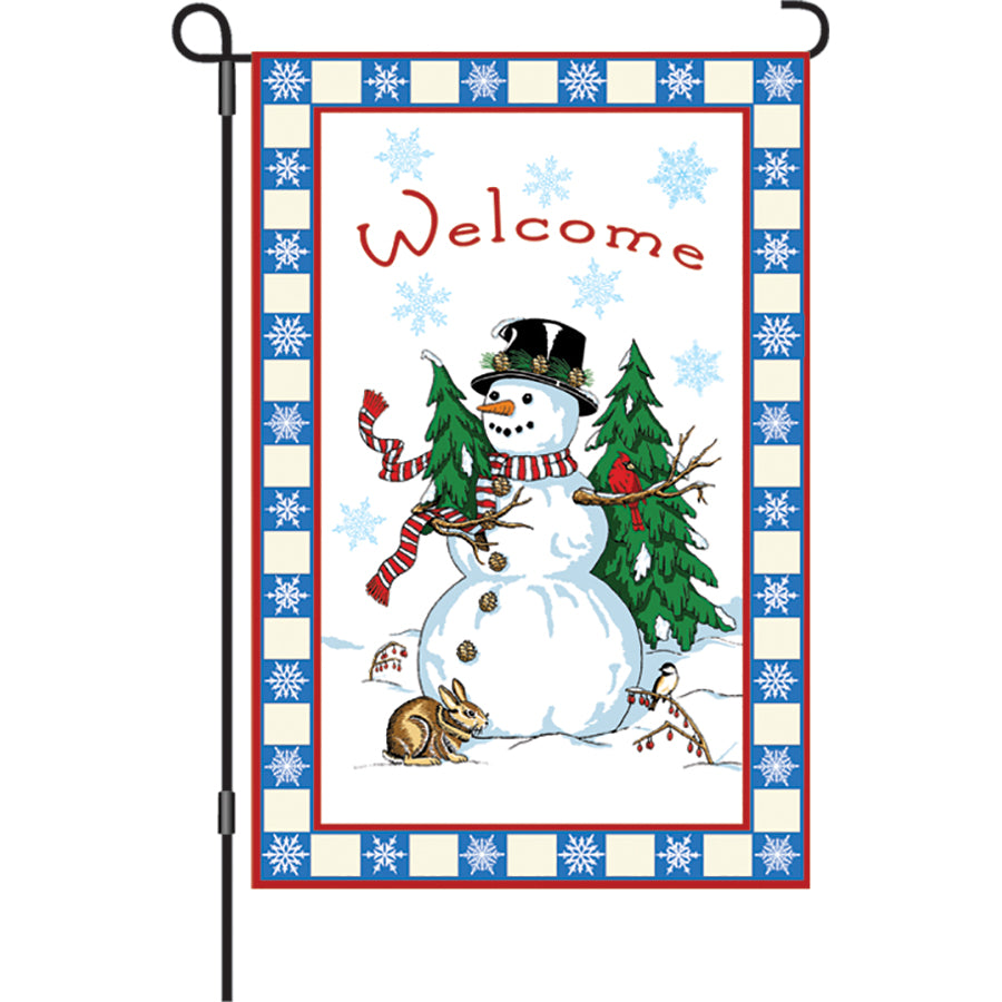 12 in. Flag - Welcome Snowman design featuring a snowman holding a Christmas tree displayed in a garden setting during winter.