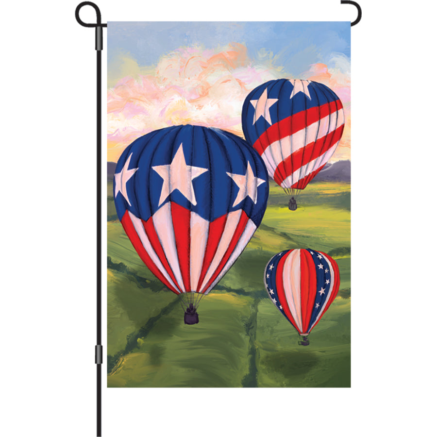 12 in. Patriotic Hot Air Balloons garden flag featuring colorful balloons against a blue sky, displayed on a flag stand in a garden setting.