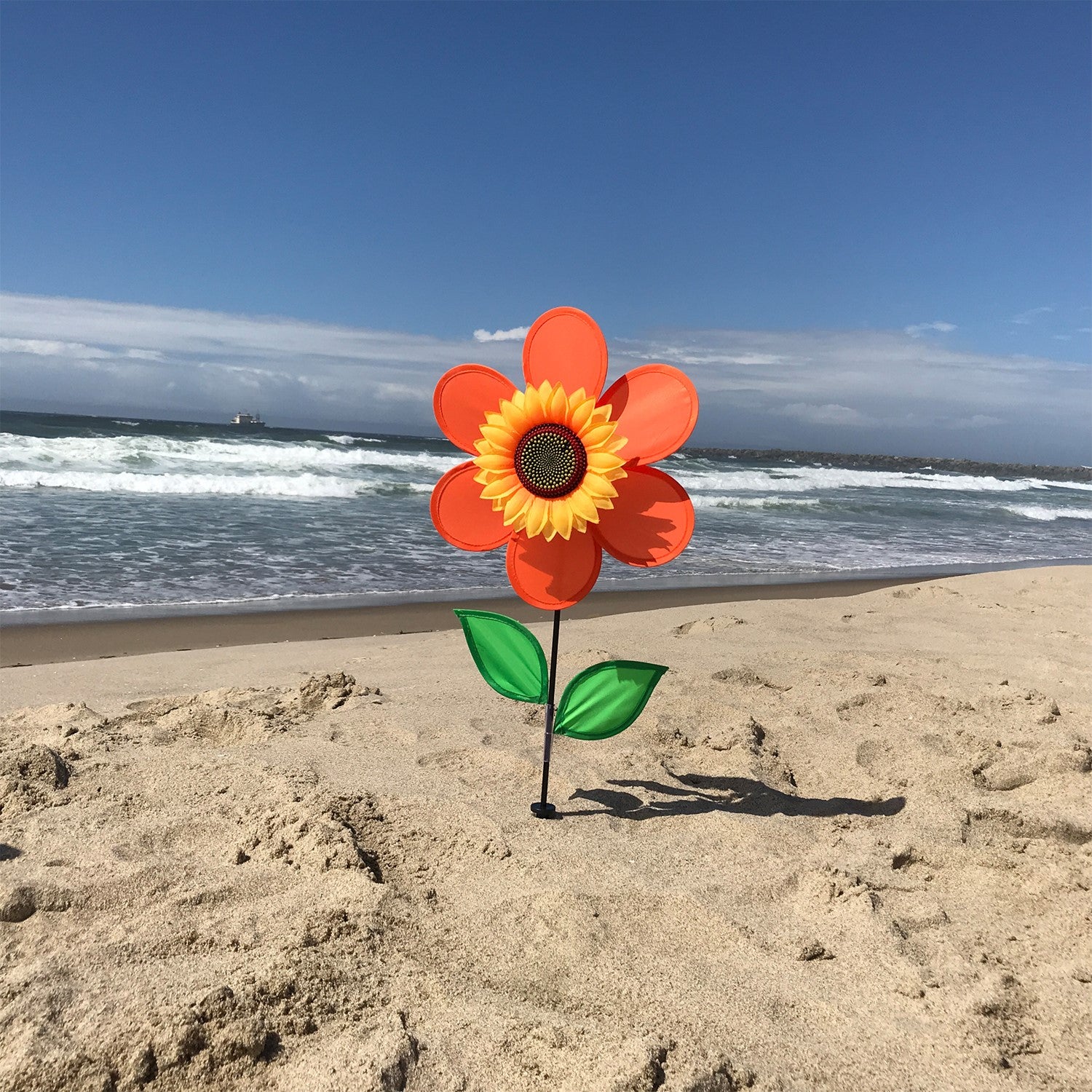12-inch orange sunflower spinner with bright petals and green leaves, spinning on a sturdy fiberglass pole in a garden setting.