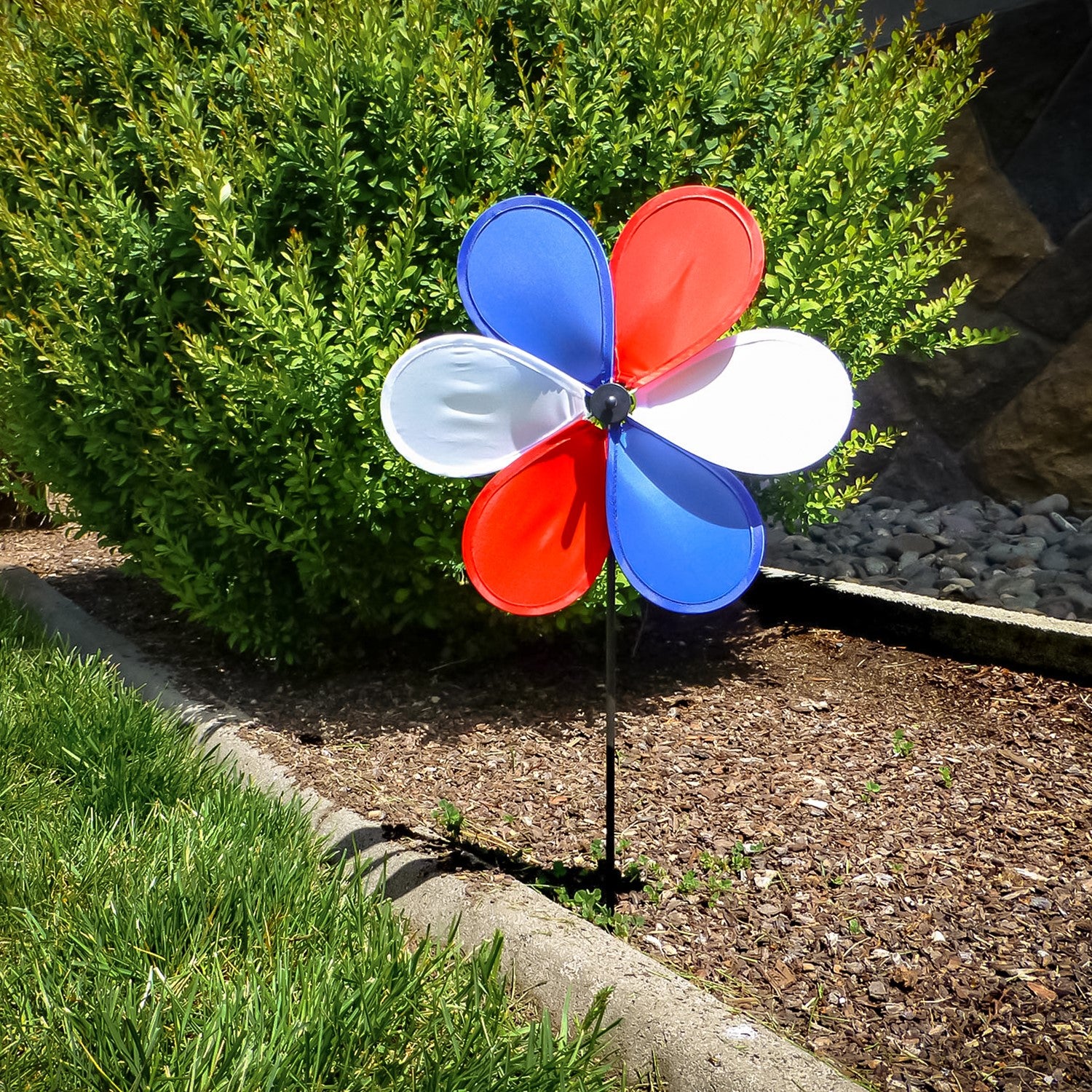 12-inch Patriot Flower spinner with red, white, and blue petals spinning on a 17-inch fiberglass pole in a garden setting.