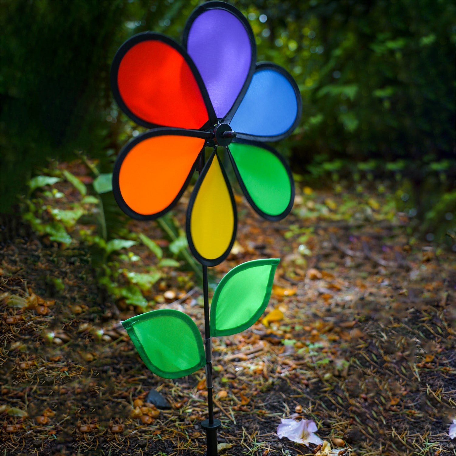12-inch Rainbow Flower Spinner with six colorful petals and leaves on a fiberglass pole, spinning in the breeze in a garden setting.
