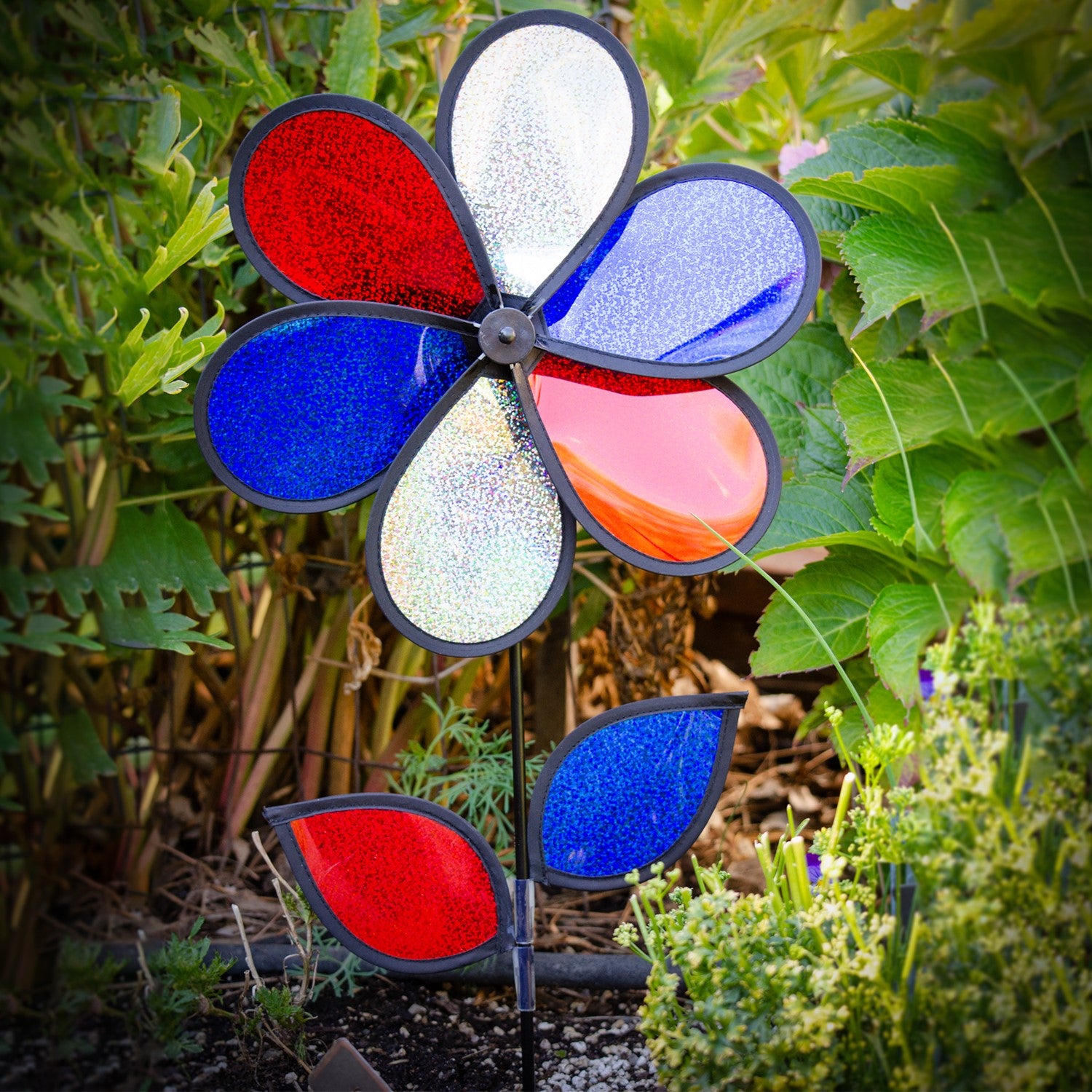 12-inch red, white, and blue sparkle flower spinner rotating in a garden, showcasing its Mylar petals and sturdy fiberglass pole.