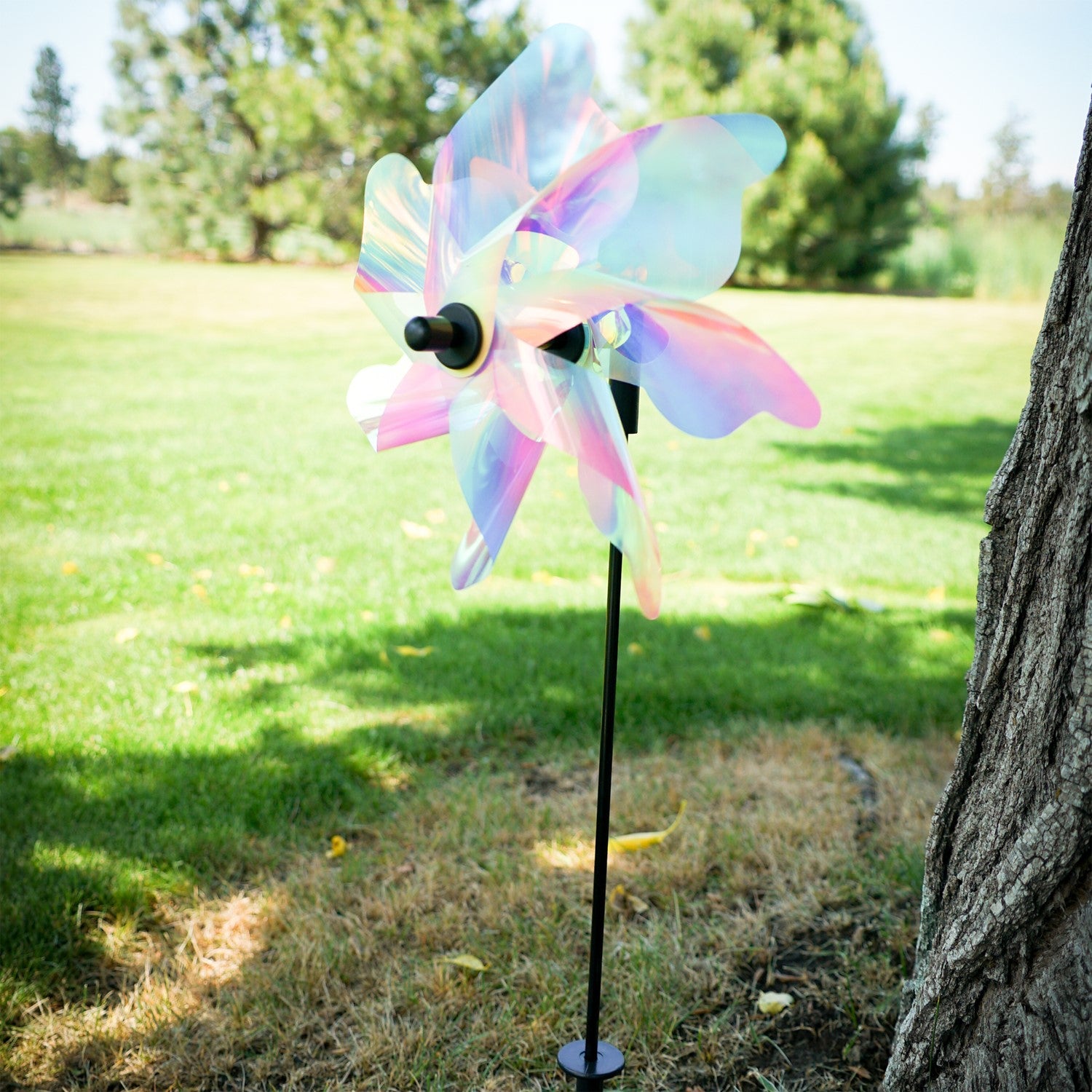 12-inch Striped Iridescent Pinwheel Spinner with pastel striped Mylar petals, displayed on a fiberglass pole in a blooming flower garden.