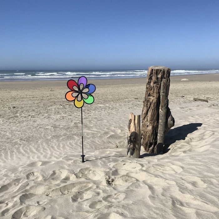 Colorful 12" Rainbow Windee Wheelz Spinner with petal design, set against a bright blue sky in a garden setting, showcasing its vibrant hues.