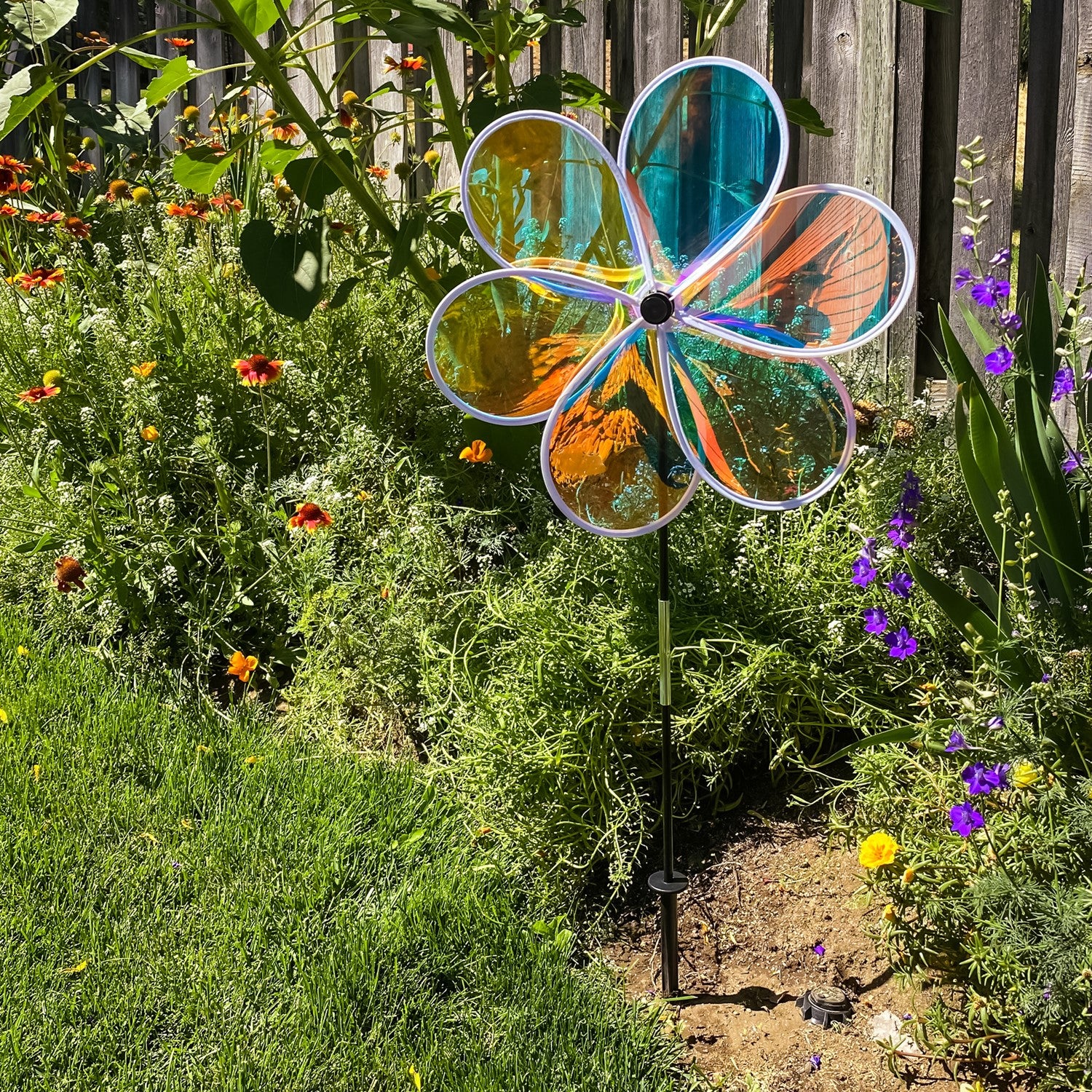 19-inch Iridescent Flower Spinner with transparent Mylar petals captured in a garden setting, reflecting sunlight and colors beautifully.