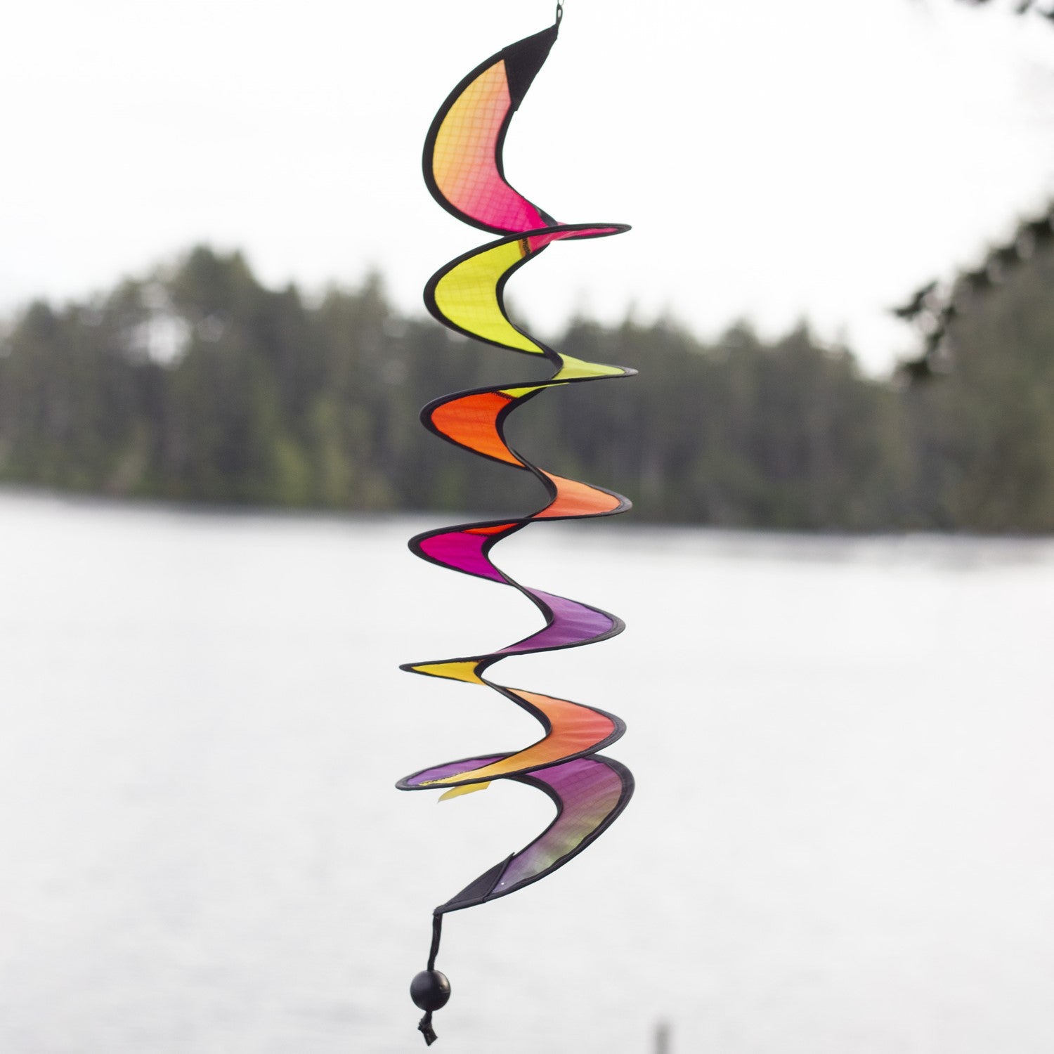 24-inch Jewel Curlie featuring a colorful rainbow graphic, hanging elegantly in an outdoor garden setting, moving gently in the breeze.