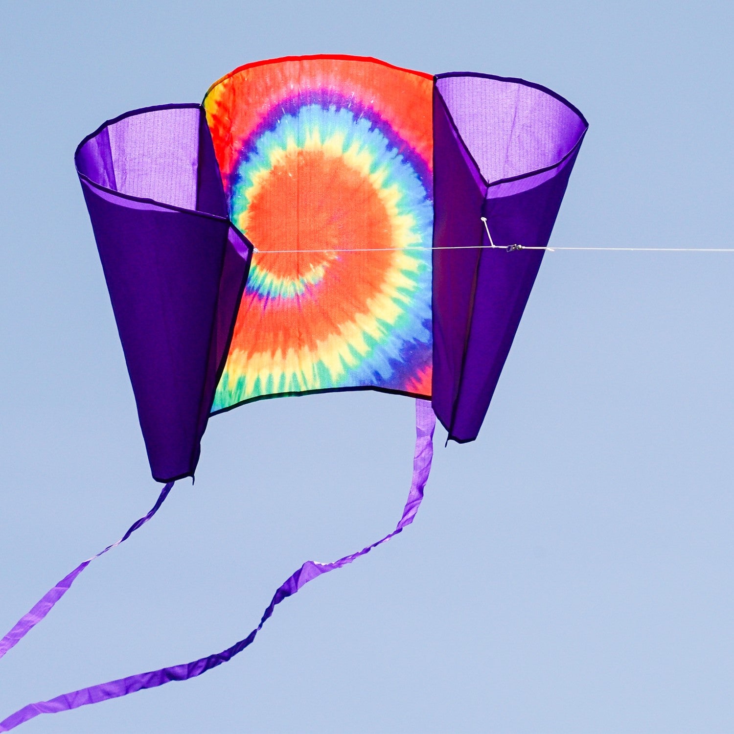 30-inch Tie Dye Sled Kite with purple tails soaring in a clear blue sky, showcasing its vibrant colors and user-friendly design for outdoor fun.