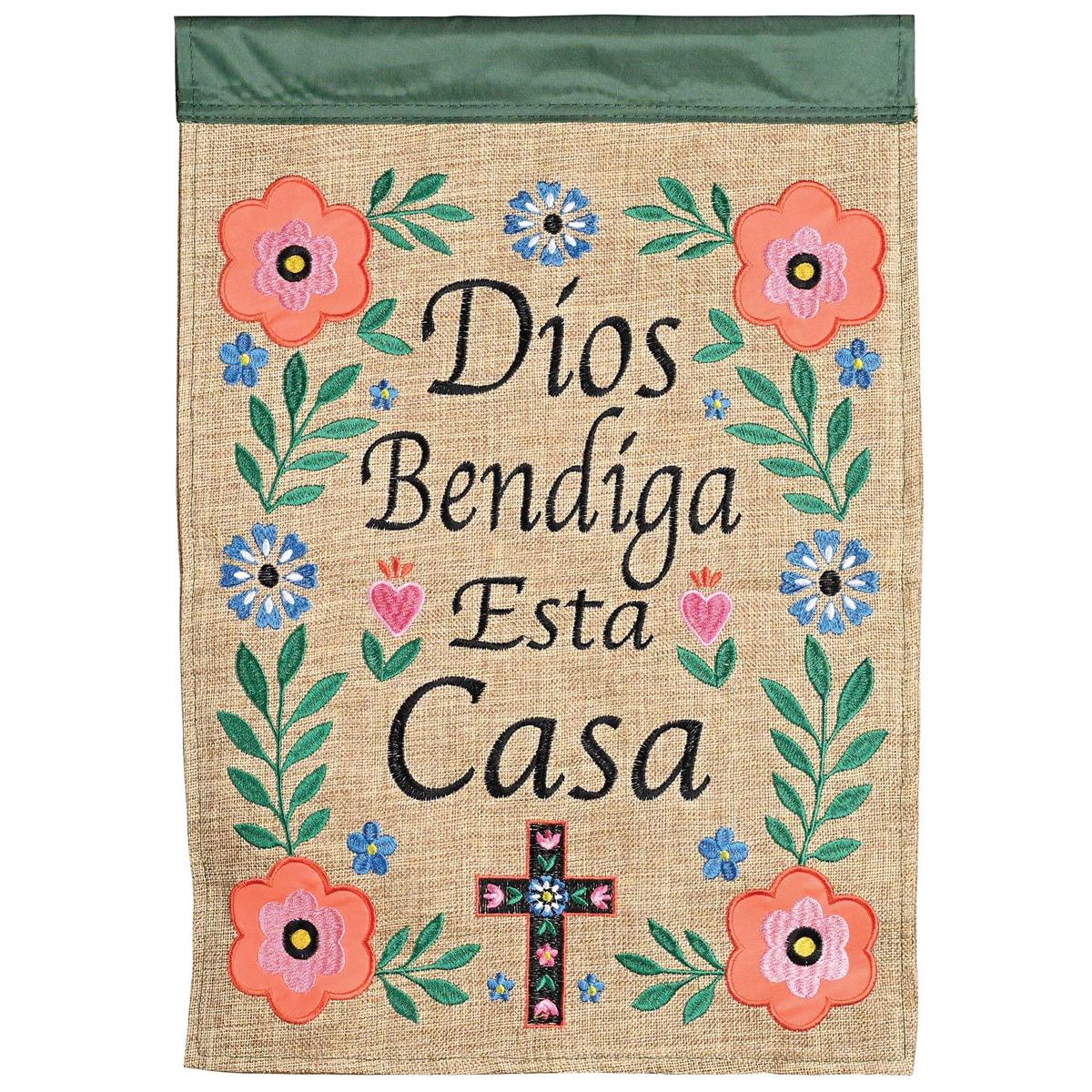 Dios Bendiga Esta Casa double-sided garden flag with intricate fabric appliques and embroidery displayed outdoors in a garden setting.