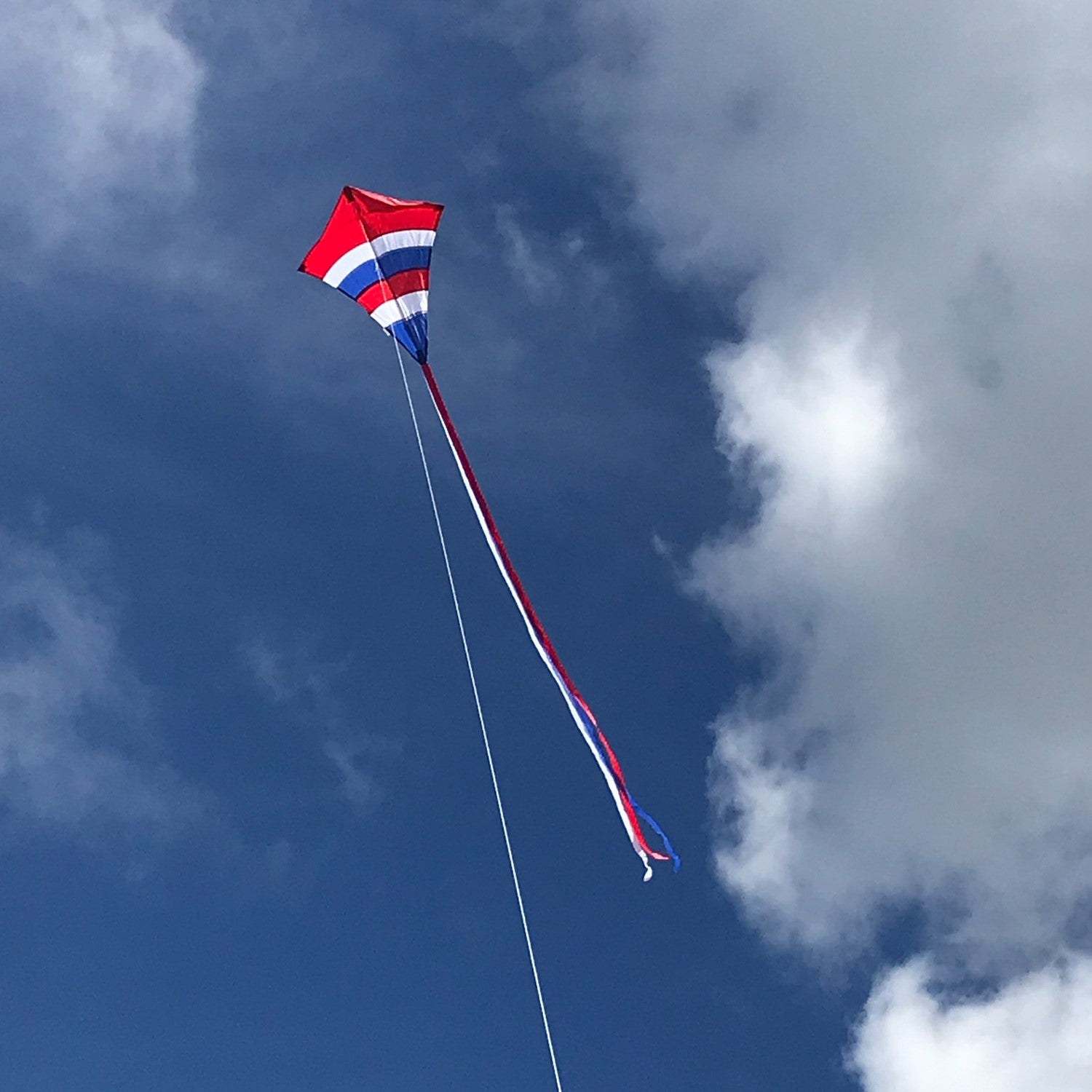 Alt Text: Patriotic Arch 27" Diamond Kite with vibrant colors and long tails flying against a blue sky during an outdoor family activity.