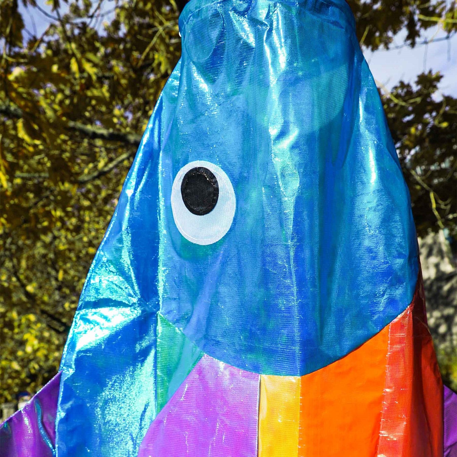 48-Inch Iridescent Rainbow Fishy Windsock featuring shimmering rainbow colors, purple fins, and a blue head, hanging in an outdoor garden.