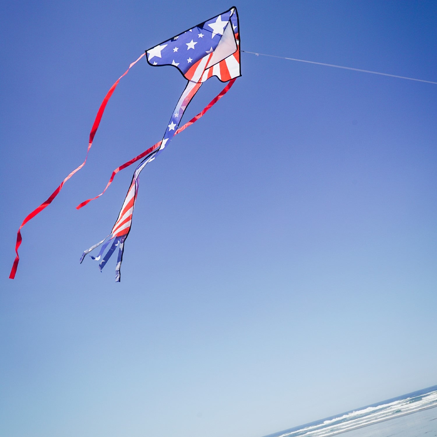 Patriotic 30-inch Mini Fly-Hi kite displaying bright stars and stripes design flying in the sky with attached 60-inch streamer tails.