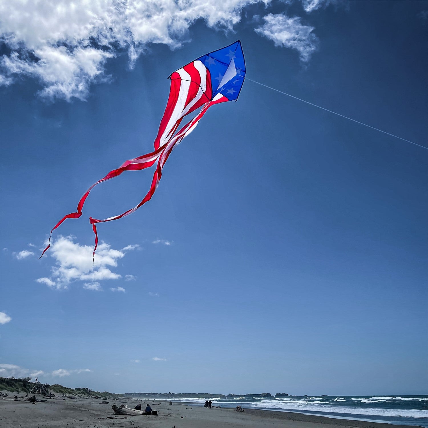 Patriotic 56" Delta kite featuring red, white, and blue stars and stripes with long tails, soaring in bright blue sky against a backdrop of fluffy clouds.