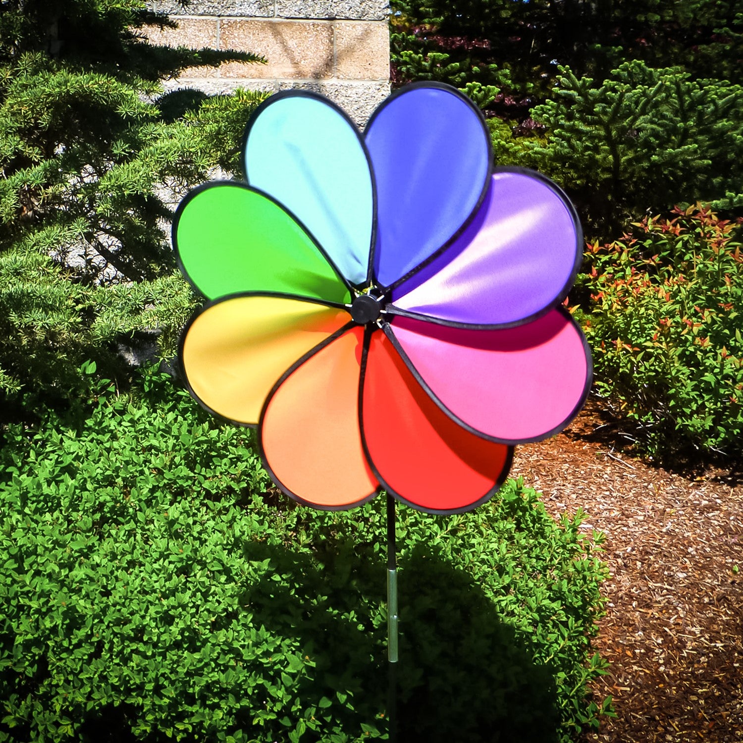 Rainbow Dazy Petal Spinner features eight colorful petals arranged in a flower design, spinning in the breeze atop a sturdy pole in a garden setting.