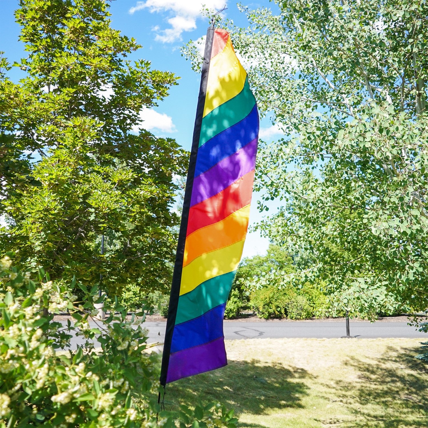 7-foot Rainbow Feather Banner with colorful applique stripes standing on an outdoor pole, illuminated by sunlight for a vibrant display.