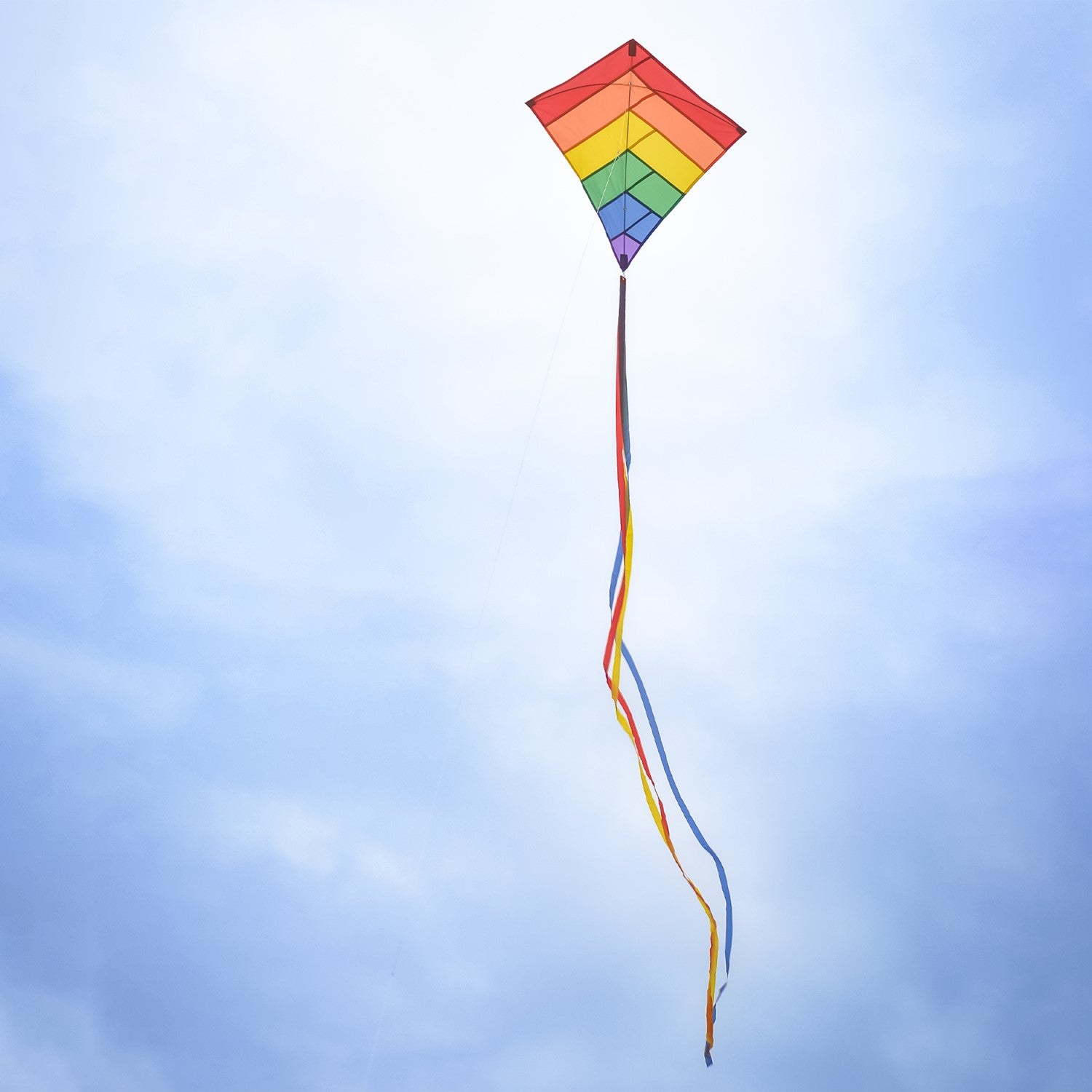 Rainbow Overlay 30-inch Diamond Kite flying in the blue sky with colorful streamer tails trailing below, showcasing its vibrant design.