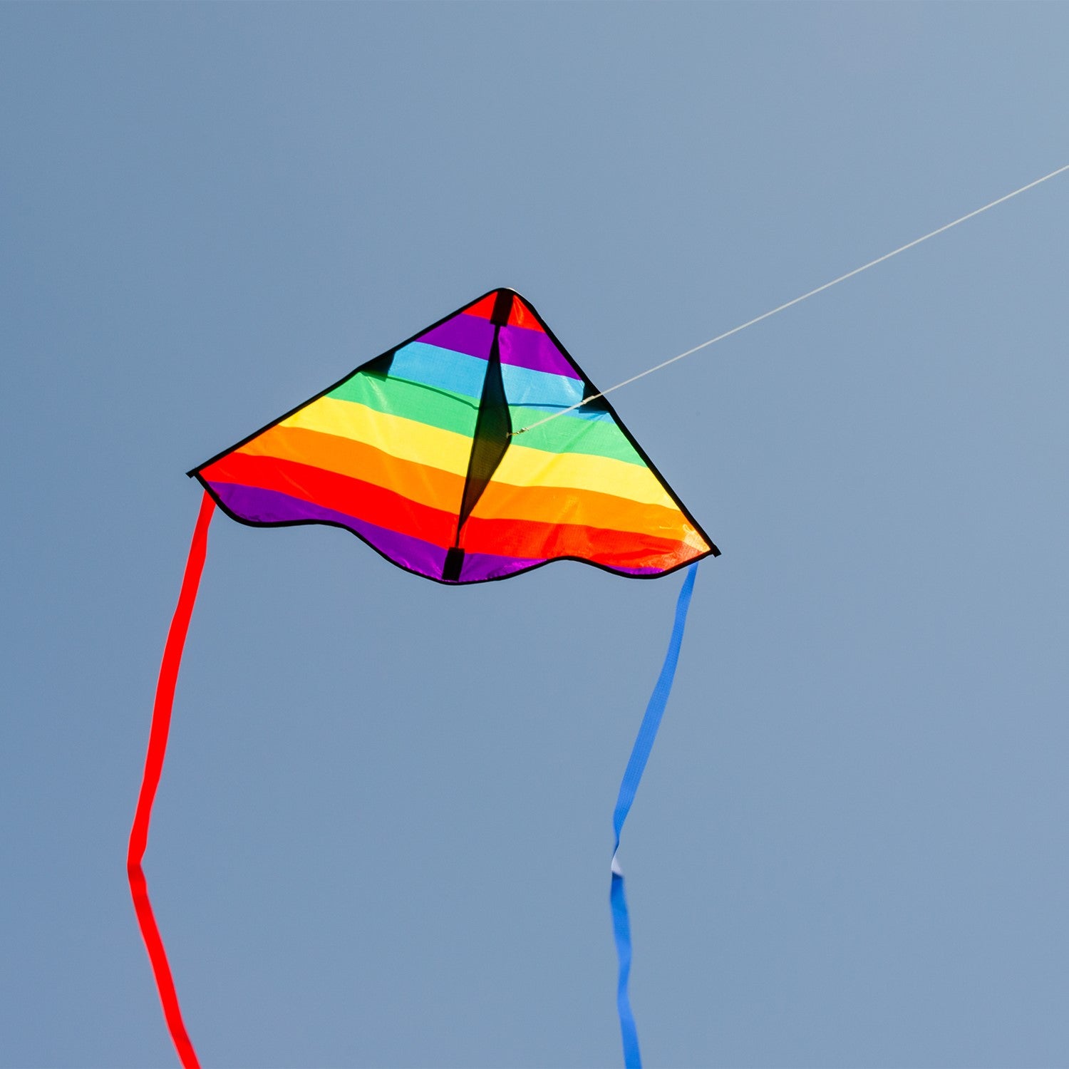 Colorful Rainbow Stripe 30-inch Delta Kite, featuring durable ripstop fabric and stable design, displayed soaring in a bright blue sky with playful tails fluttering below.
