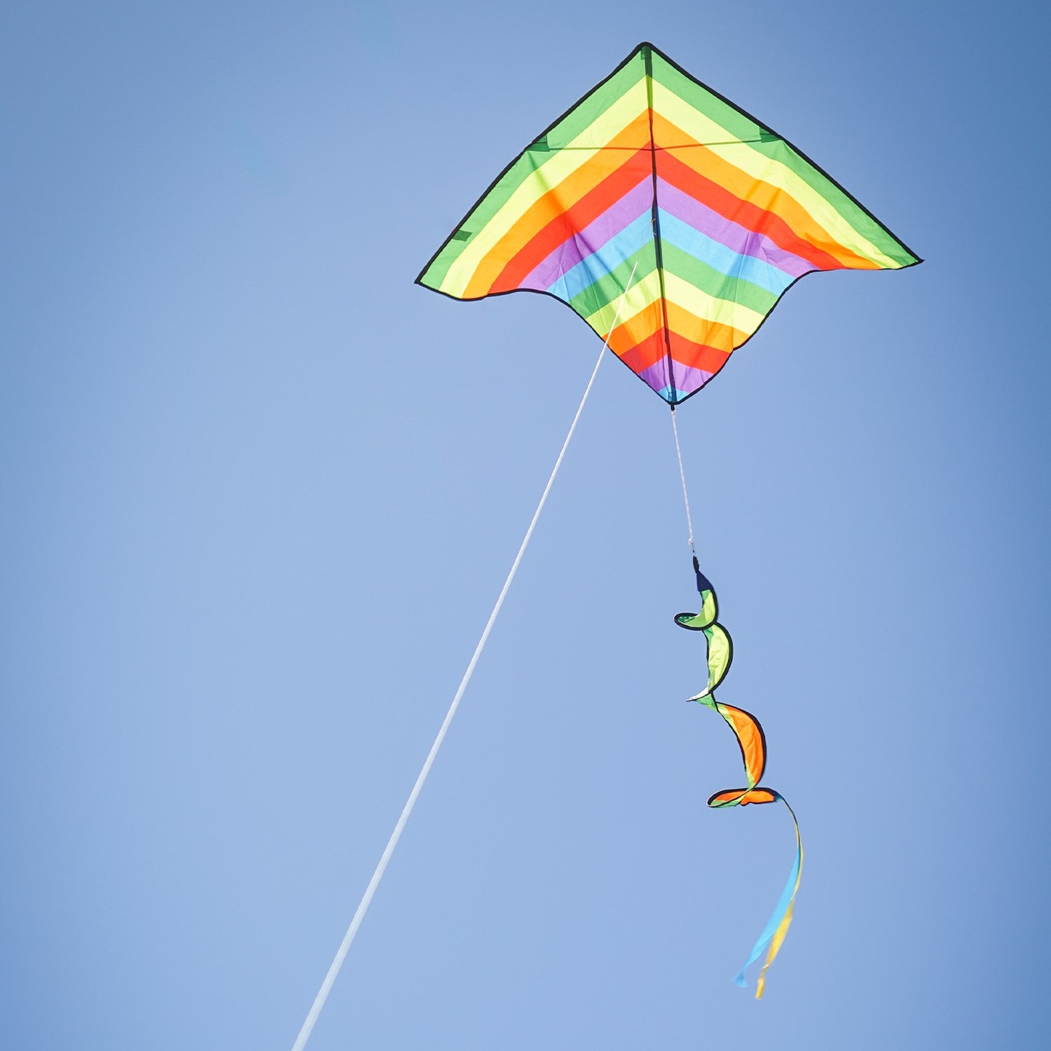 Rainbow Stripe Delta kite with a curlie spinning tail flying against a clear blue sky and green grass backdrop, showcasing its vibrant colors.