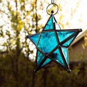 Teal 5-Point Star Hanging Tealight Lantern with textured glass panels and metal accents, suspended outdoors against a scenic backdrop.