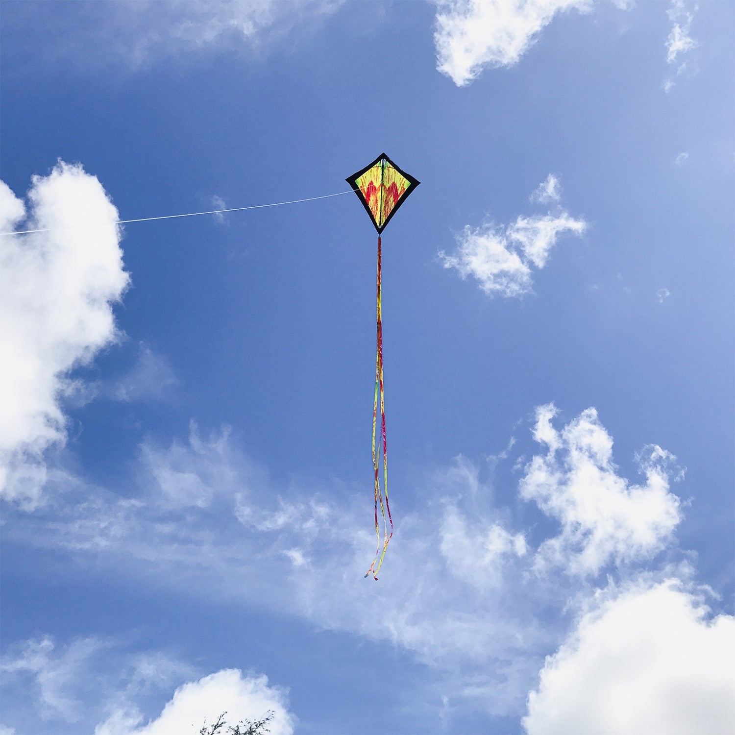 Tie Dye 30 inch Diamond Kite flying high in the sky with vibrant colors and 12-foot streamer tails swaying in the wind.