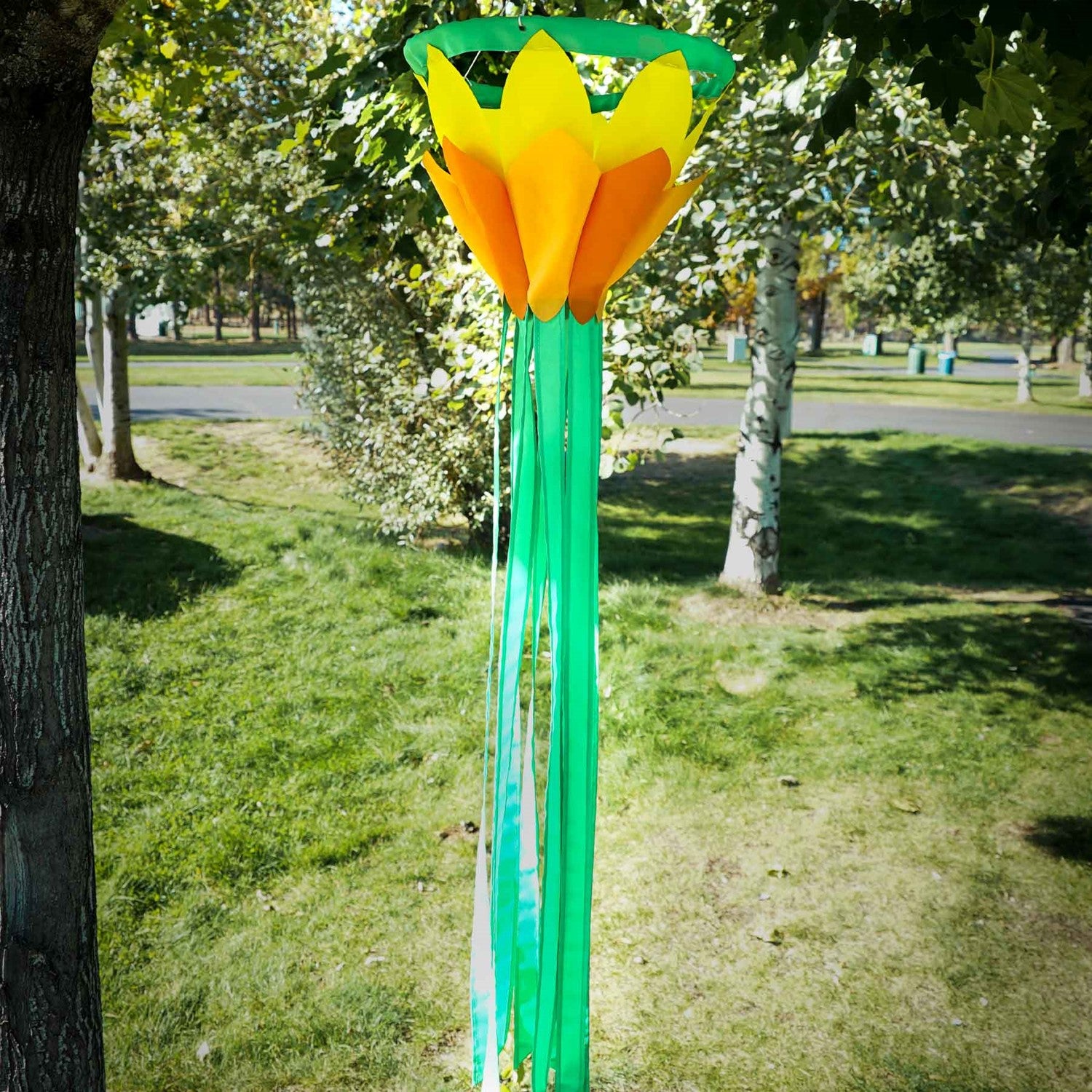 Colorful Yellow 38-Inch Lotus Flower Windsock featuring vibrant yellow and orange petals with green tails, displayed outdoors in a garden setting.