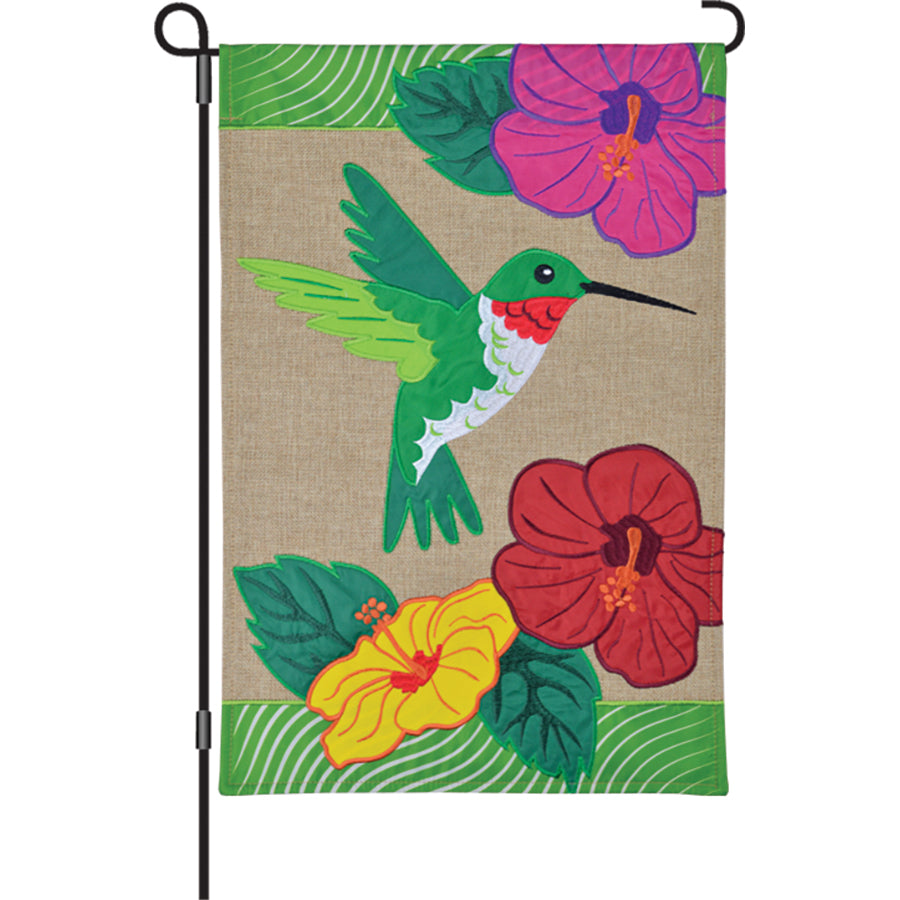 12 in. Burlap Flag with embroidered Hummingbird and Hibiscus design displayed in a garden setting, adding charm and color to the outdoor area.