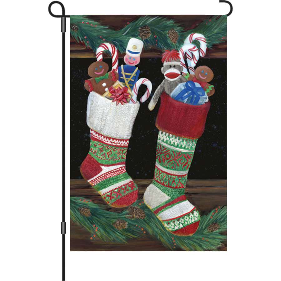 12 in. Christmas Stocking garden flag featuring vibrant festive designs, displayed outdoors in a winter-themed garden setting.
