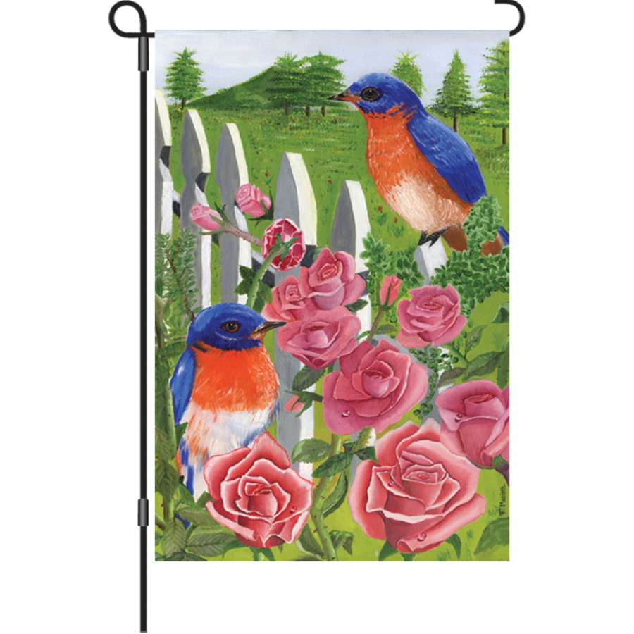 12 in. illuminated Bluebird & Rose garden flag featuring a bluebird on roses displayed outdoors in a vibrant garden setting.