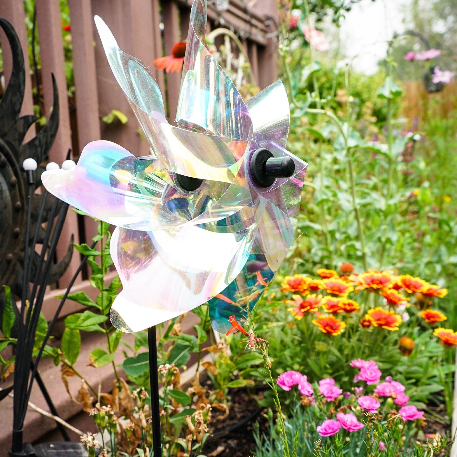 12-inch iridescent pinwheel spinner displayed in a garden, featuring colorful Mylar petals that catch the wind and reflect sunlight.