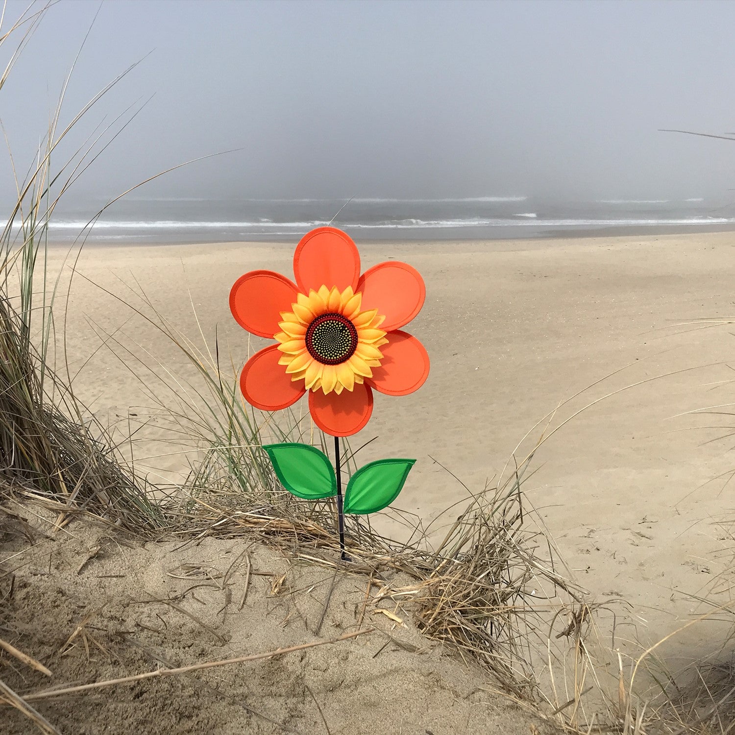 12-inch orange sunflower spinner with bright petals and green leaves, spinning on a sturdy fiberglass pole in a garden setting.
