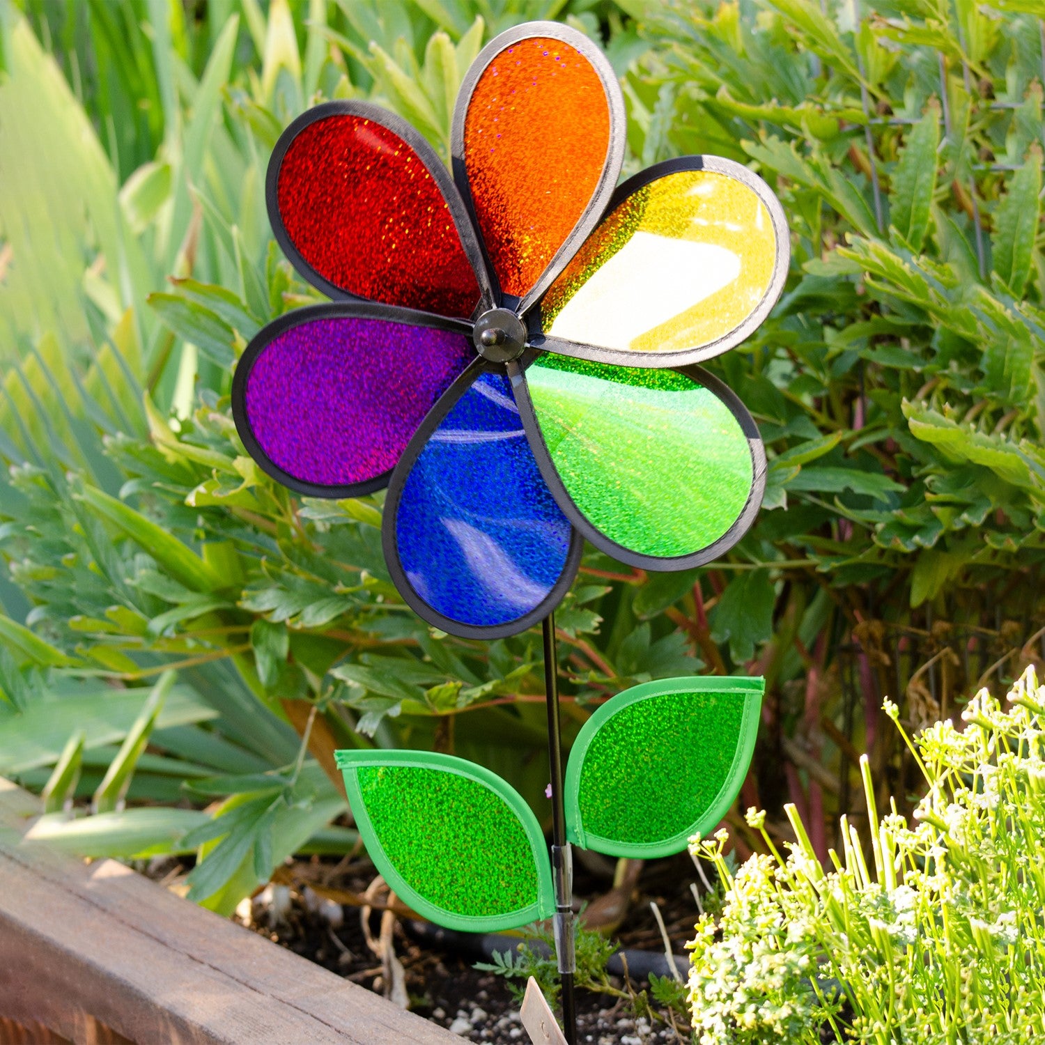 12-inch Rainbow Sparkle Flower Spinner featuring six colorful Mylar petals and green leaves on a fiberglass pole in a garden setting.