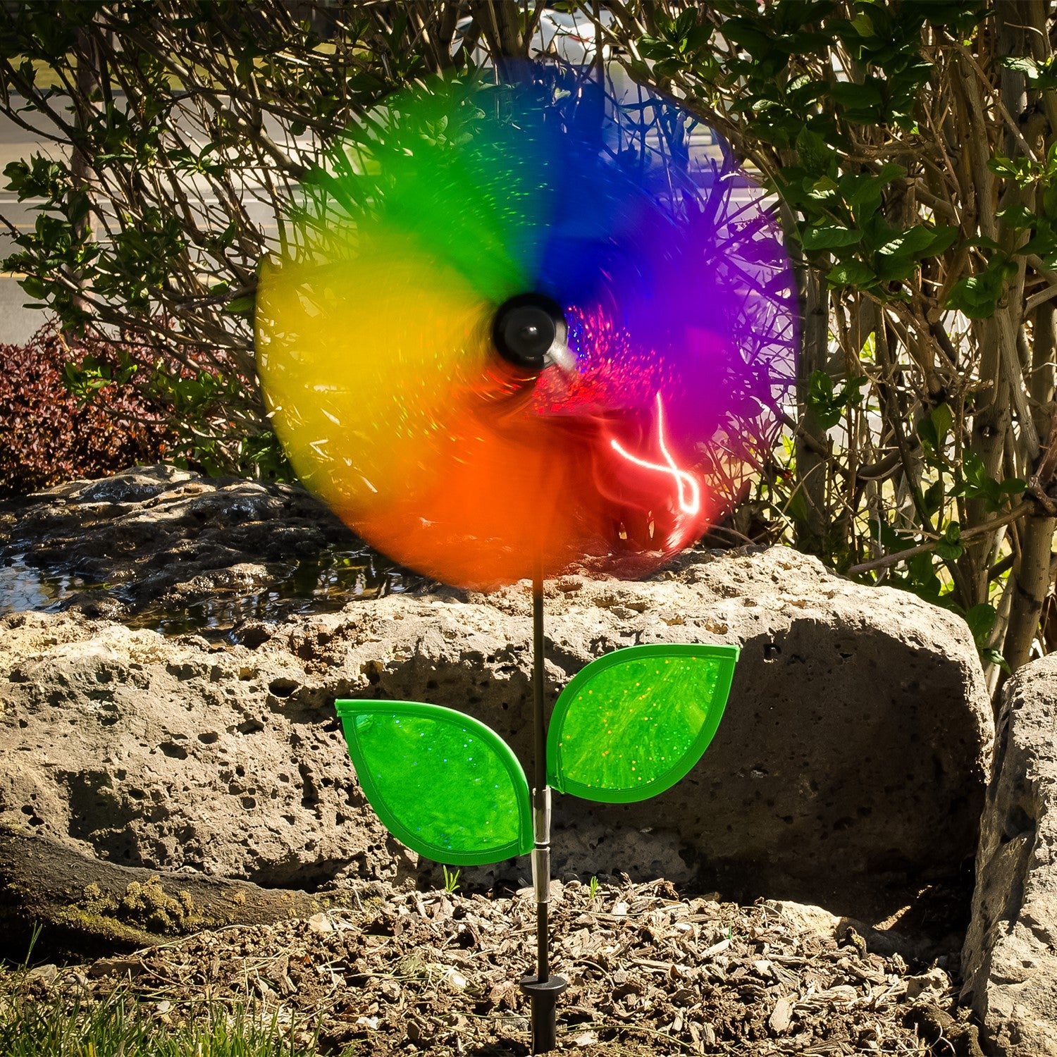 12-inch Rainbow Sparkle Flower Spinner featuring six colorful Mylar petals and green leaves on a fiberglass pole in a garden setting.