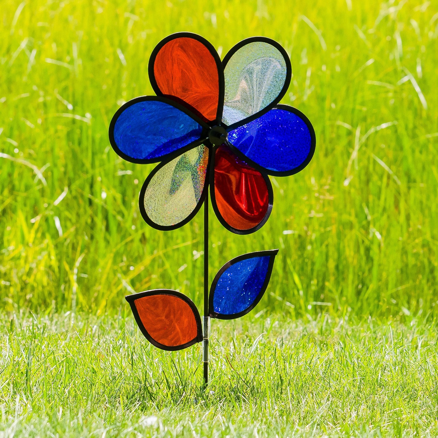 12-inch red, white, and blue sparkle flower spinner rotating in a garden, showcasing its Mylar petals and sturdy fiberglass pole.