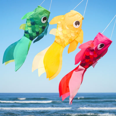 Three colorful Damsel Fish windsocks in pink, yellow, and green, hanging in a garden, gently moving with the breeze under a sunny sky.