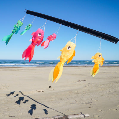 Three colorful Damsel Fish windsocks in pink, yellow, and green, hanging in a garden, gently moving with the breeze under a sunny sky.