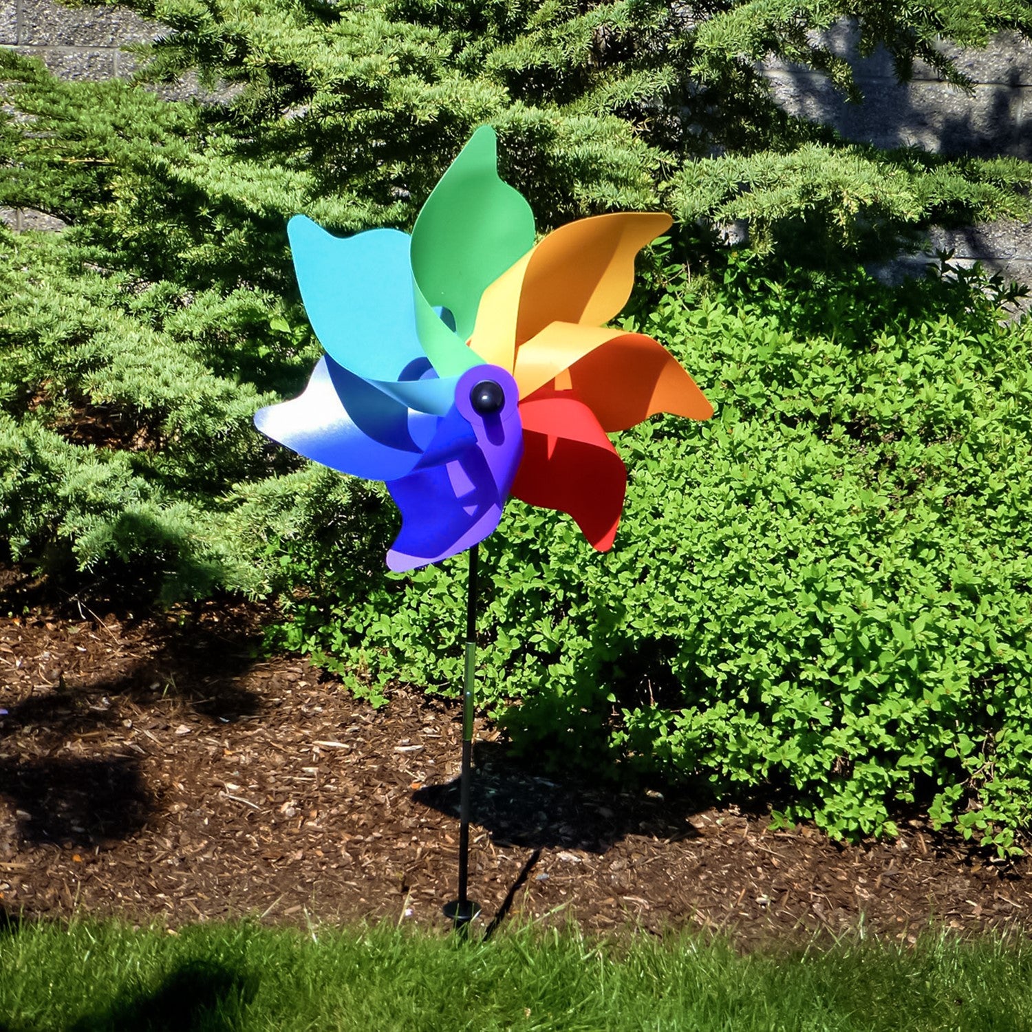 A vibrant 18\" Rainbow Poly Petal Spinner showing colorful petals spinning in the breeze, mounted on a two-section fiberglass pole in a garden setting.