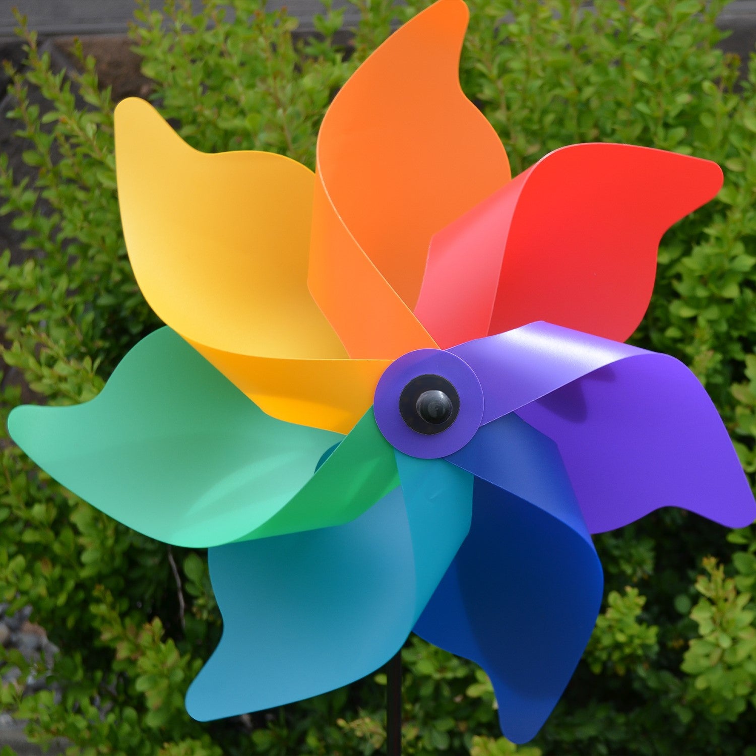 A vibrant 18\" Rainbow Poly Petal Spinner showing colorful petals spinning in the breeze, mounted on a two-section fiberglass pole in a garden setting.