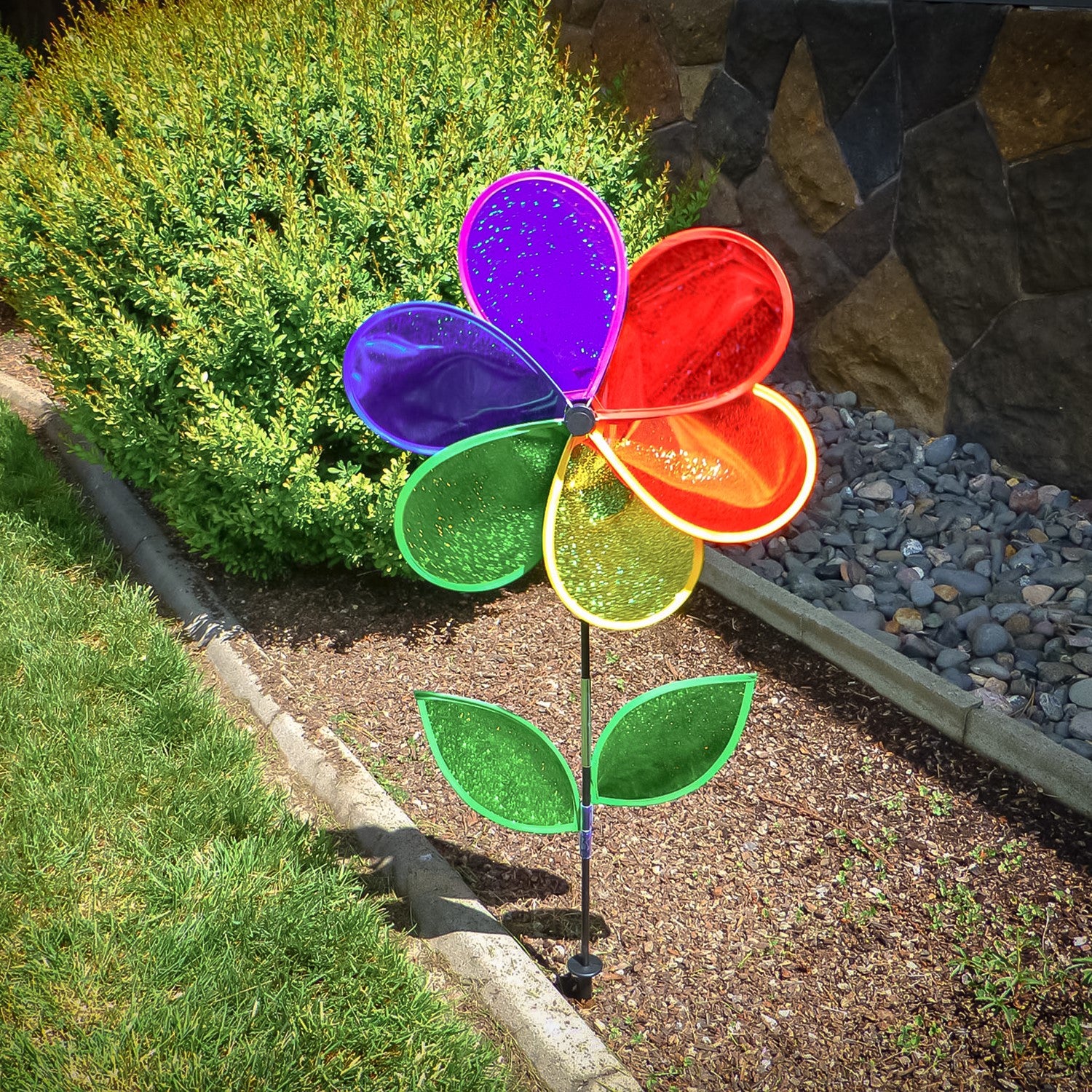19 inch Rainbow Glitter Flower Spinner with bright Mylar petals spinning on a garden pole in a sunny outdoor yard setting.