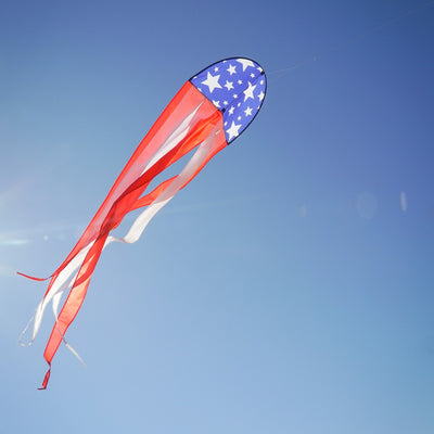 22" Patriot Dancing Dragon Kite with stars and stripes design soaring through the sky during a sunny outdoor event with tails flowing.