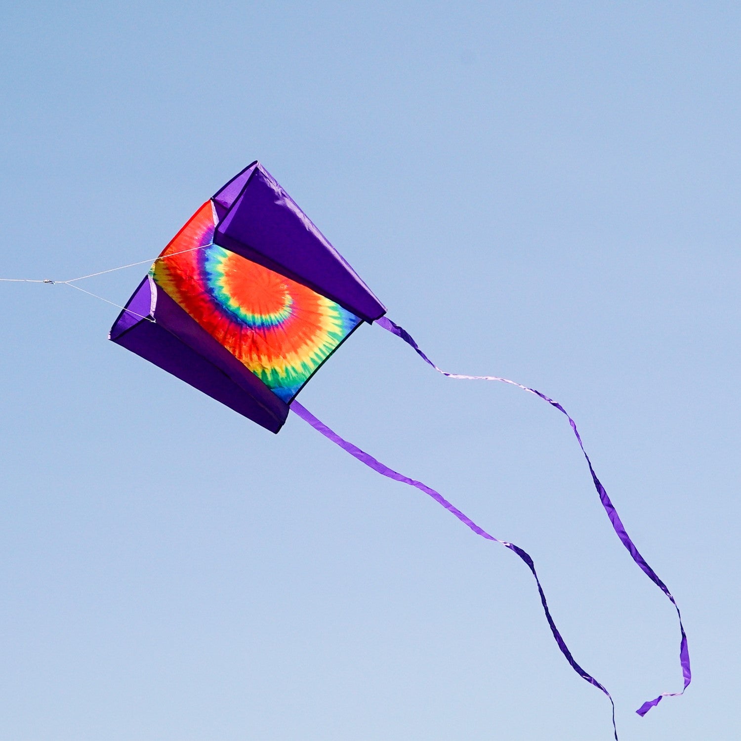 30-inch Tie Dye Sled Kite with purple tails soaring in a clear blue sky, showcasing its vibrant colors and user-friendly design for outdoor fun.