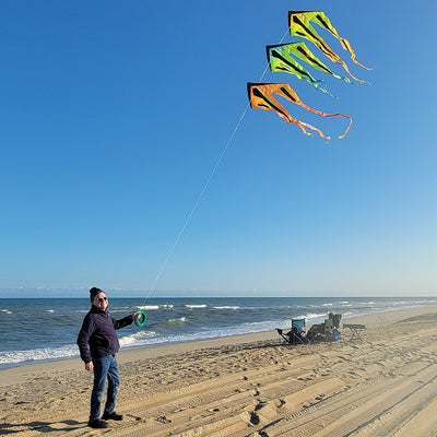46 in. Flo-Train Delta Kite in vibrant mango design with long 10-foot tail flying against a blue sky in an outdoor setting.
