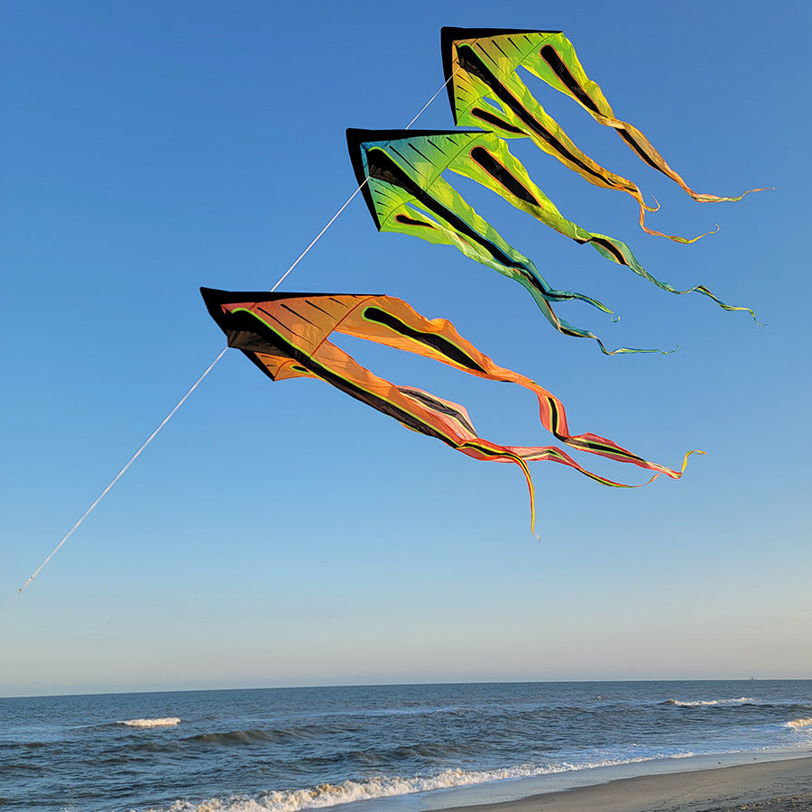 46 in. Flo-Train Delta Kite - Orange Shock Flag soaring against a clear blue sky, displaying its long colorful tails in an outdoor setting.