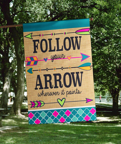 Rustic Follow Your Arrow burlap banner flag 29x42 with double applique arrows and bold lettering, shown hanging on a porch pole beside potted plants and a wooden bench.
