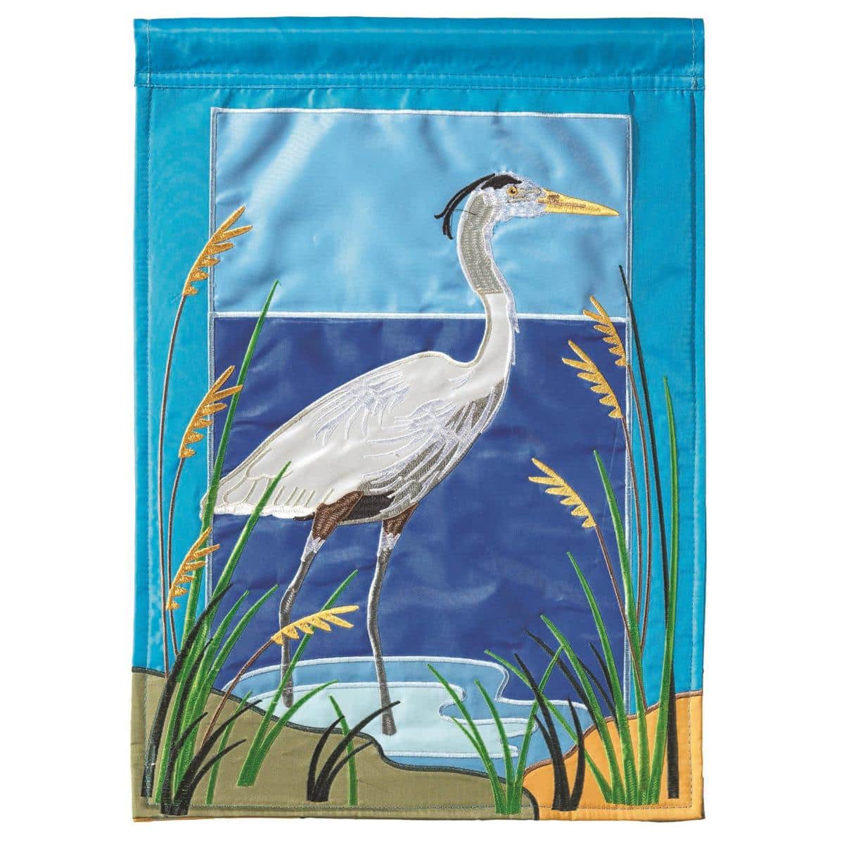 13x18 inch Heron Garden Flag showing a layered double applique heron standing by reeds with a blue water background, displayed on a garden flagpole in a landscaped yard.