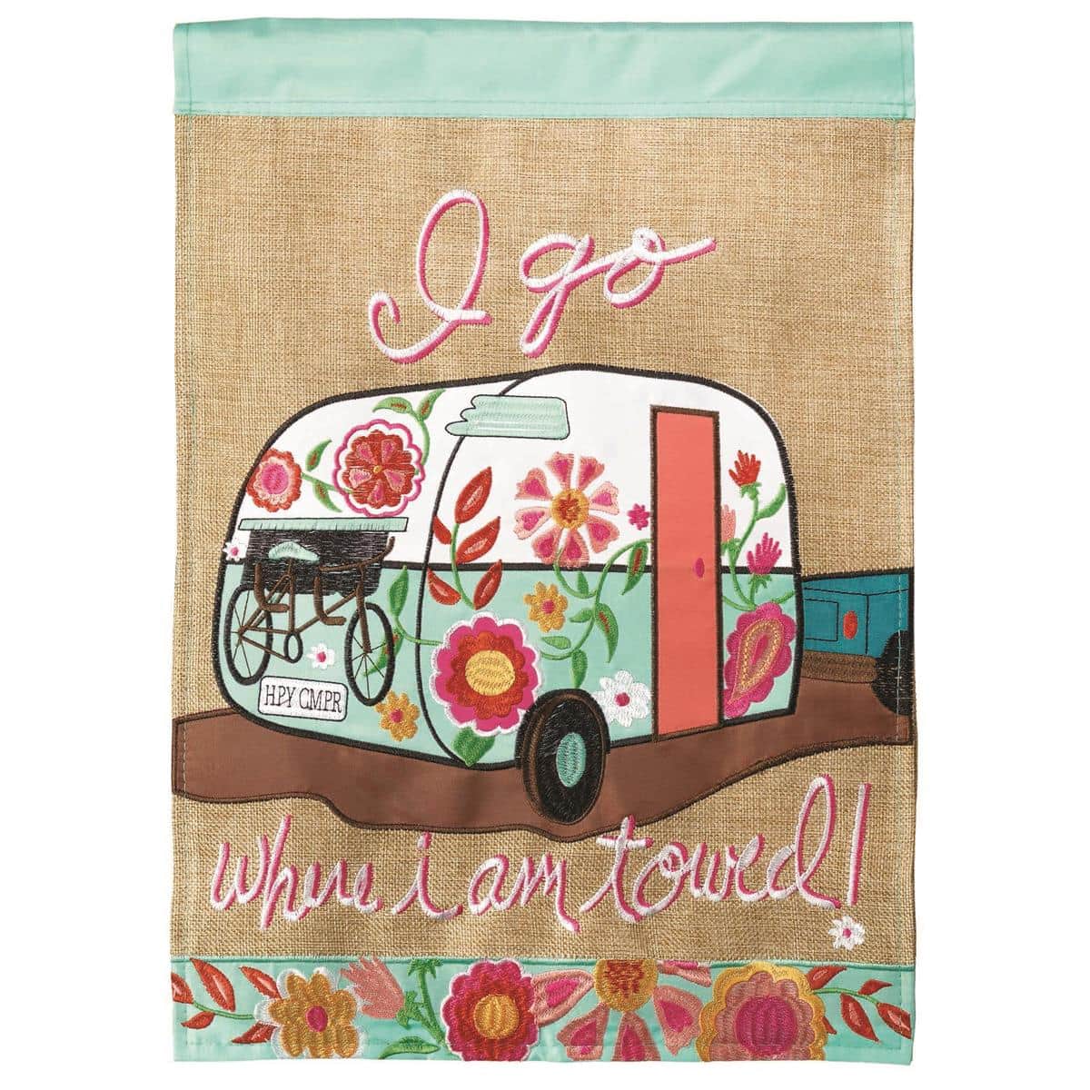Alt text: 13x18 I Go Where I Am Towed burlap garden flag with double applique camping trailer motif displayed on a garden stand beside a rustic planter.