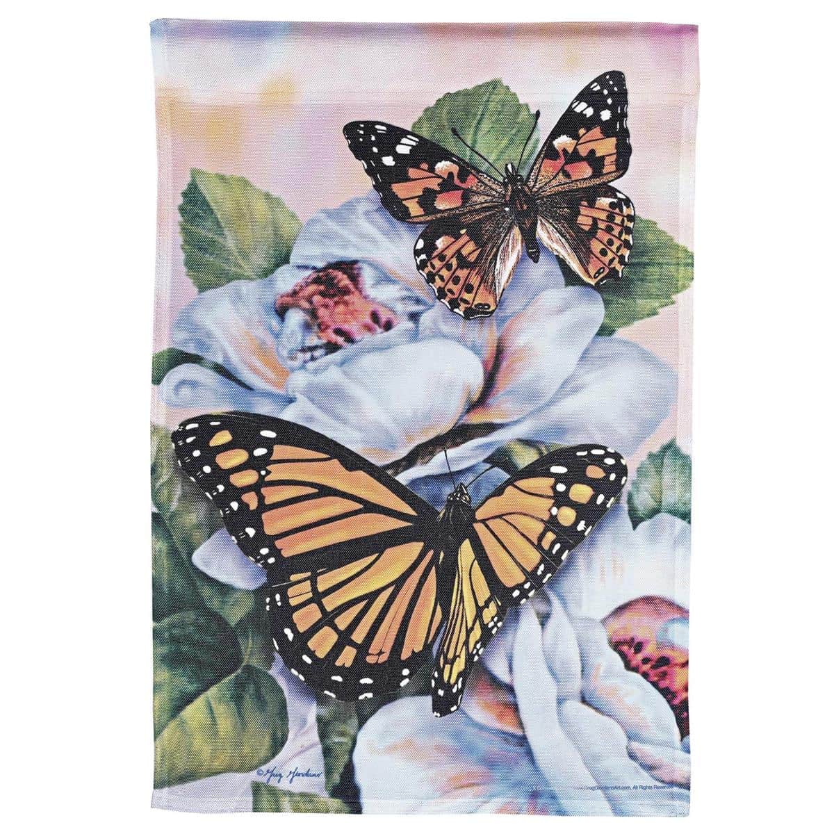 13x18 printed garden flag featuring monarch butterflies and pink and yellow floral accents hanging on a lawn flag stand beside potted plants