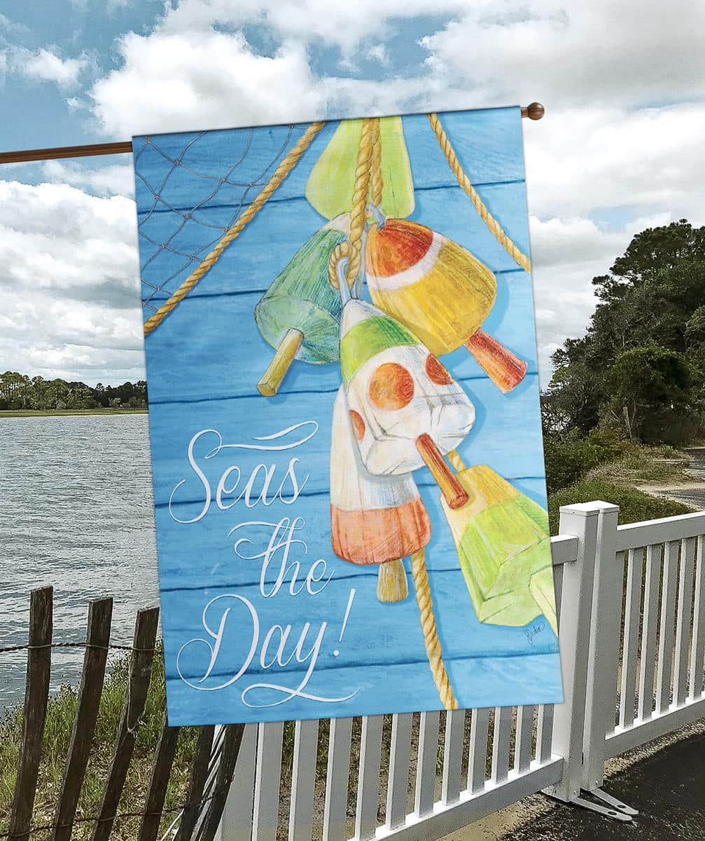 Seas The Day banner flag hanging on a front porch with navy and sand tones, showing playful 'Seas The Day' text and coastal wave and shell motifs in a printed coastal design.