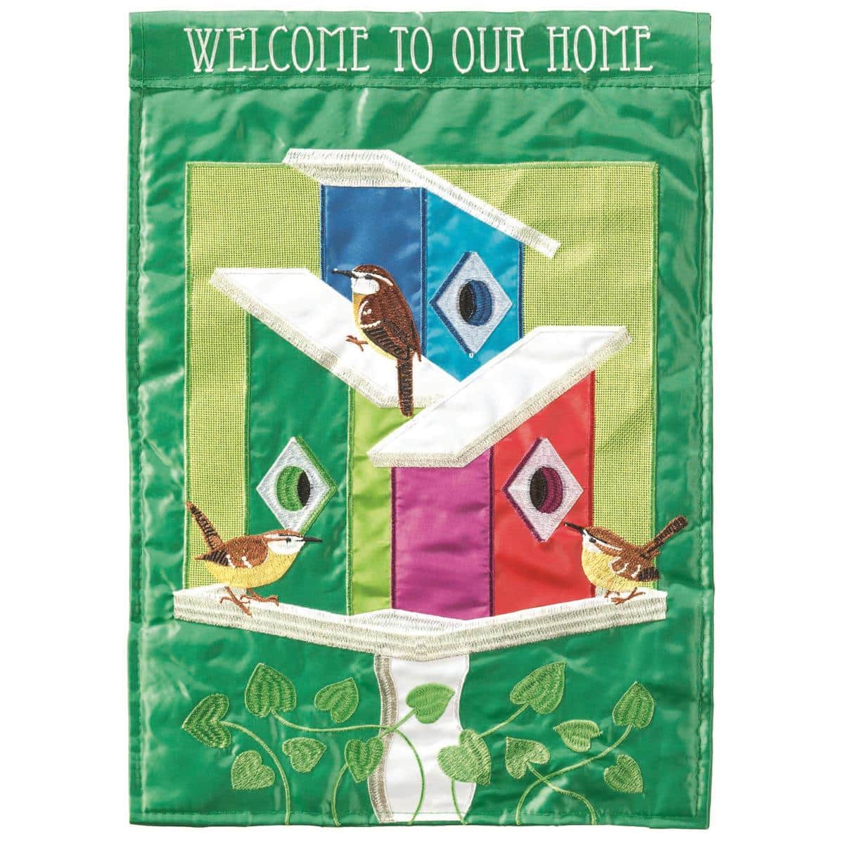 13x18 Welcome To Our Home burlap garden flag with double applique birds and birdhouse design shown hanging on a black metal yard holder near potted plants and a wooden porch step.