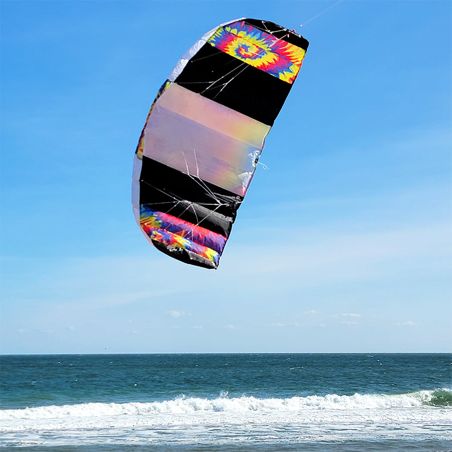 Barracuda 1.7 Kite in vibrant tie-dye pattern flying high against a blue sky, showcasing its frameless design and dual lines, perfect for outdoor fun.